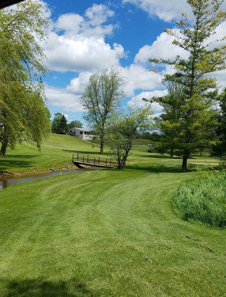 A small creek running through a golf fairway with a clubhouse sitting on a hill in the background.