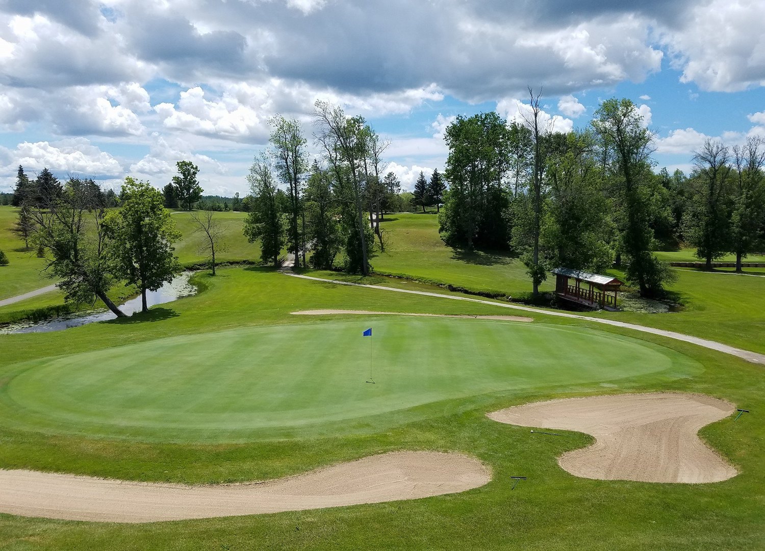 A large, wide golf green flanked by four bunkers. There is a small creek on one side of the green and a cart path and a covered foot bridge on the other. This is the 9th hole at Cheboygan Golf & Country Club.